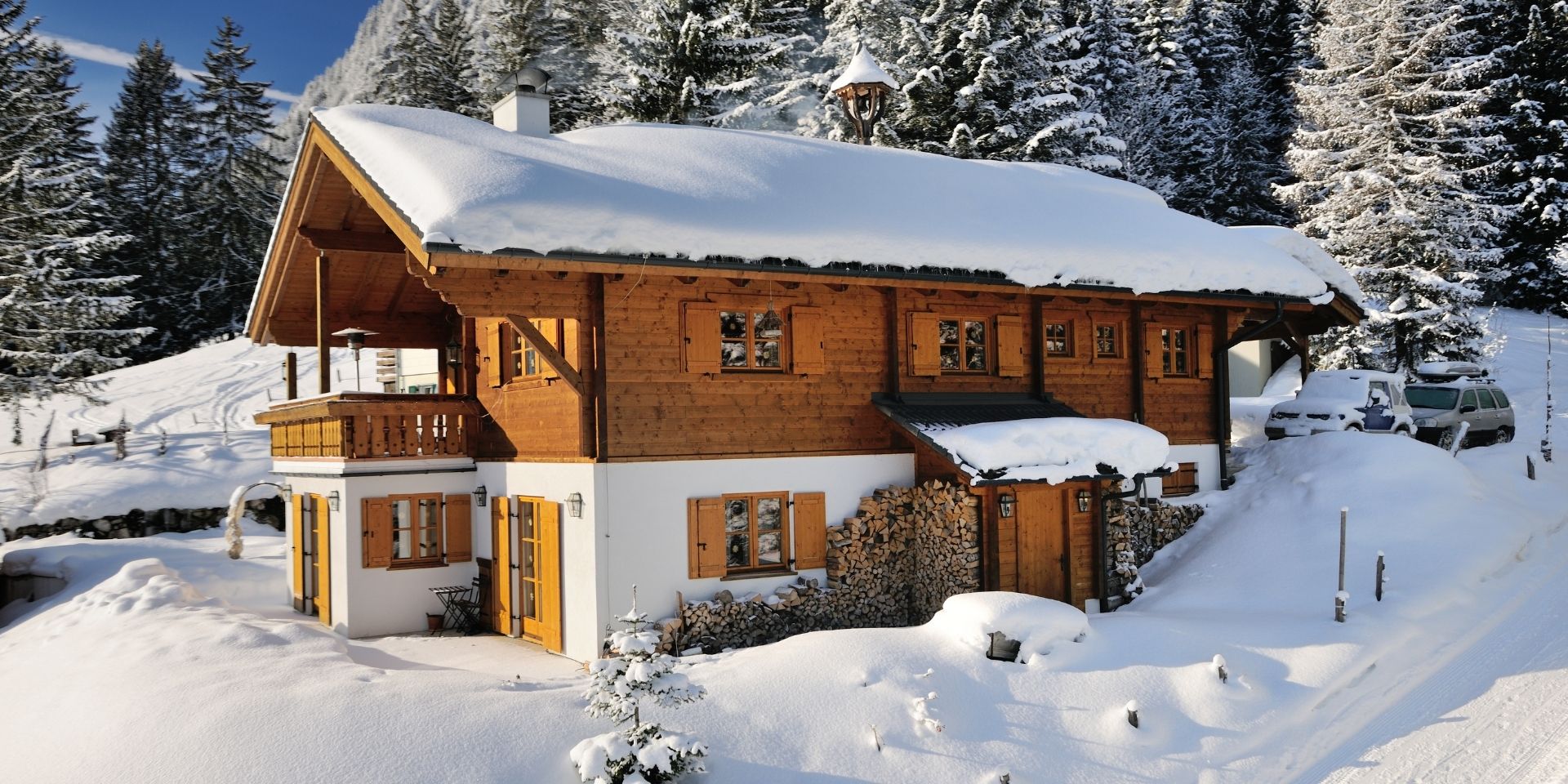 Wooden chalet covered in snow with trees and cars in the background