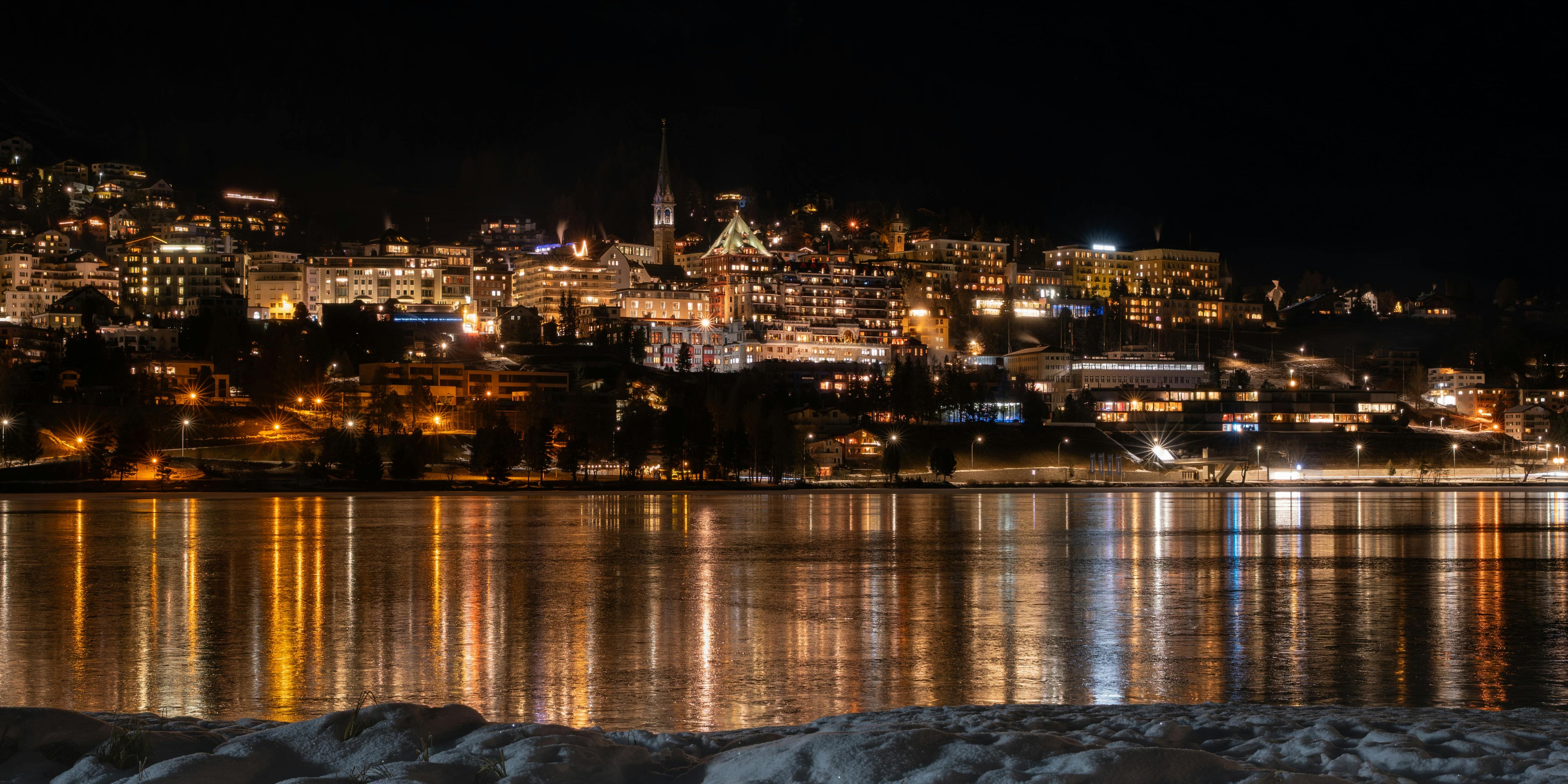 Cityscape at night with illuminated buildings reflected in a calm body of water.