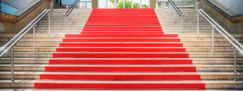 A bright red carpet runner laid up the center of a wide outdoor stone staircase with metal handrails on both sides.
