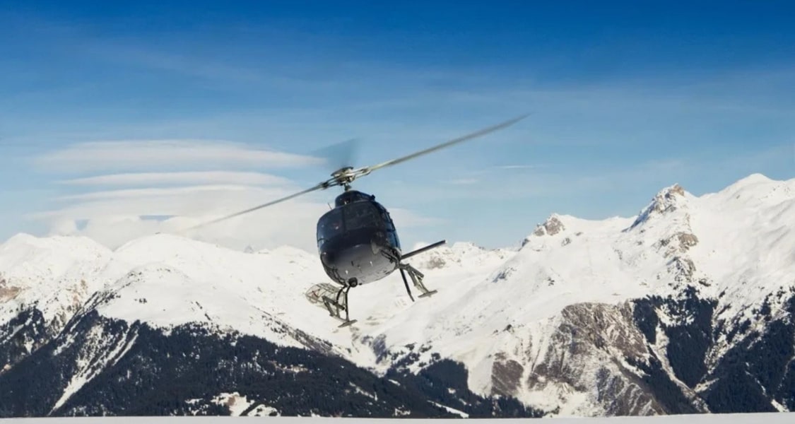 Helicopter flying over snow-covered mountains with a clear blue sky.