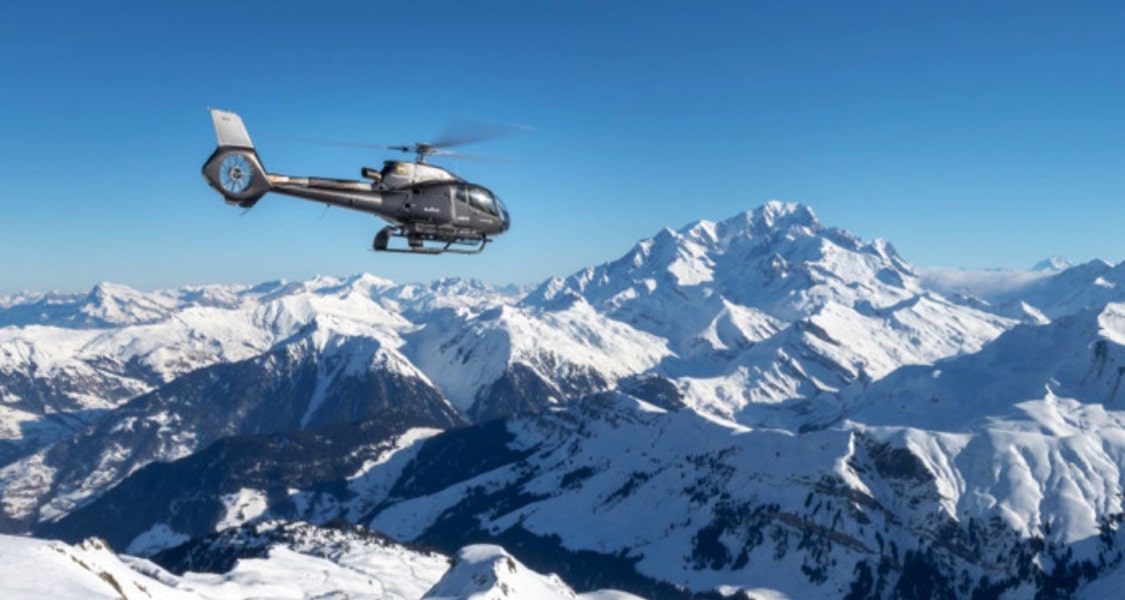 Helicopter flying over snow-covered mountains with a clear blue sky.