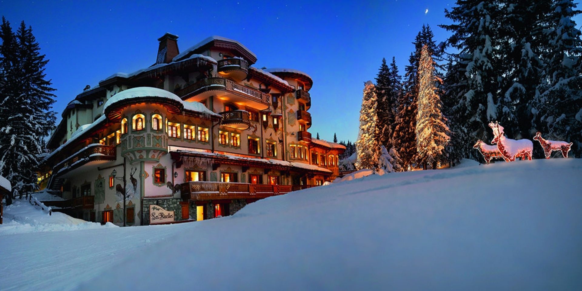 Snow-covered mountain lodge at night with illuminated windows, surrounded by trees and snow.
