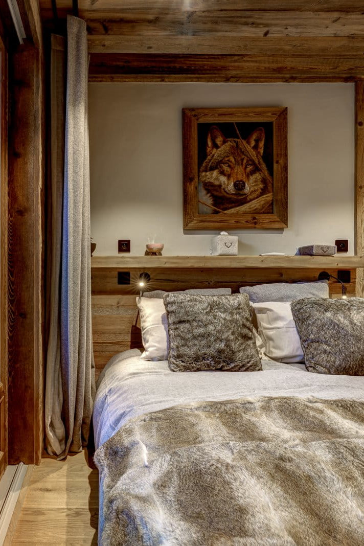 A rustic bedroom interior with a bed dressed in white and gray fur-like bedding, set in a wood-paneled cabin with a large window overlooking a garden path and trees.