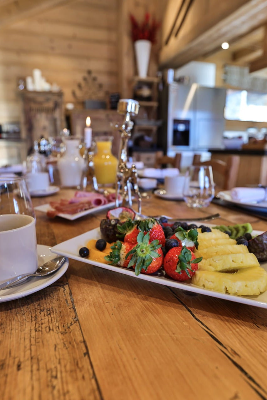 A breakfast spread on a rustic wooden table in a cabin dining area, featuring a plate with strawberries, pineapple, and sausage, alongside a white coffee cup and glasses.