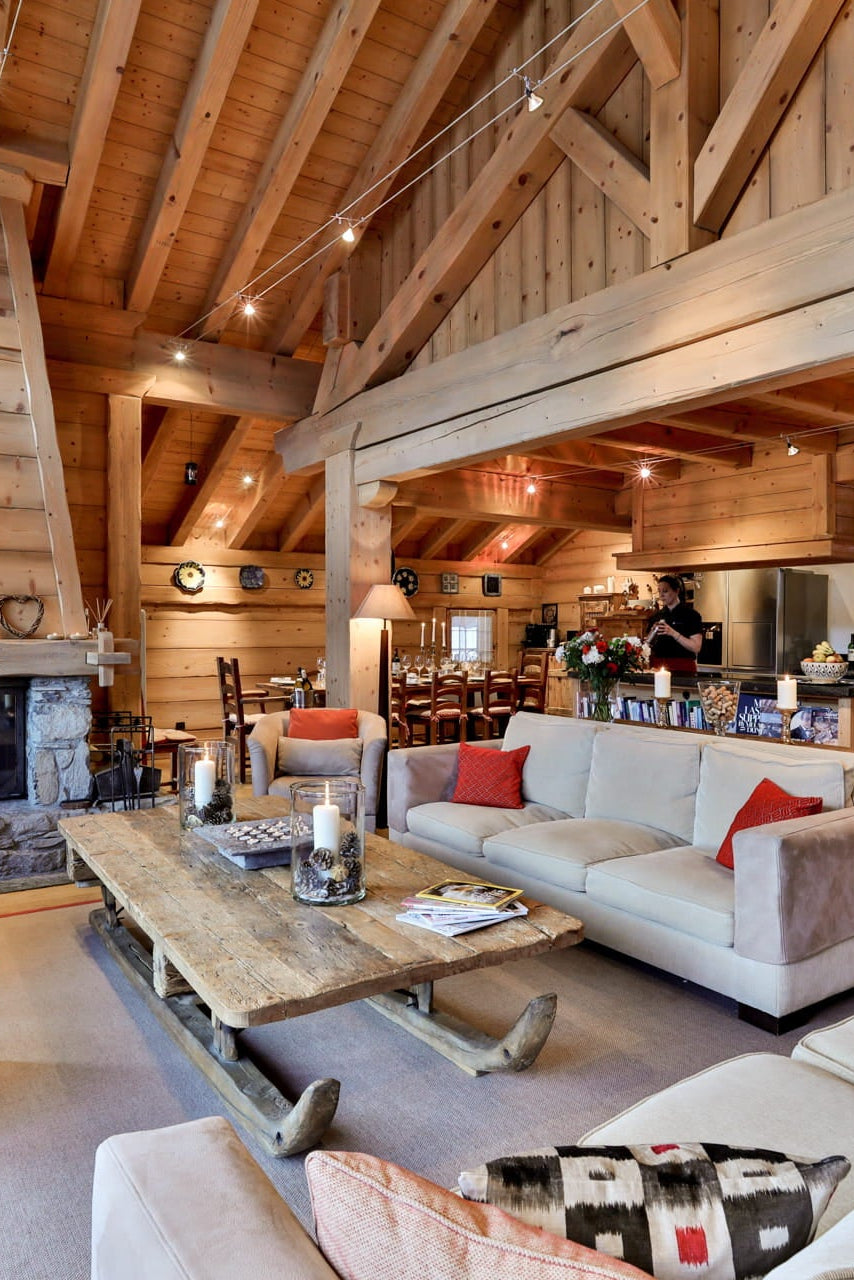 A rustic, high-ceilinged lodge-style living room with exposed wood beams, a large stone fireplace, a red armchair and ottoman, a cream sofa with red pillows, a wooden coffee table, and an open kitchen area in the background.