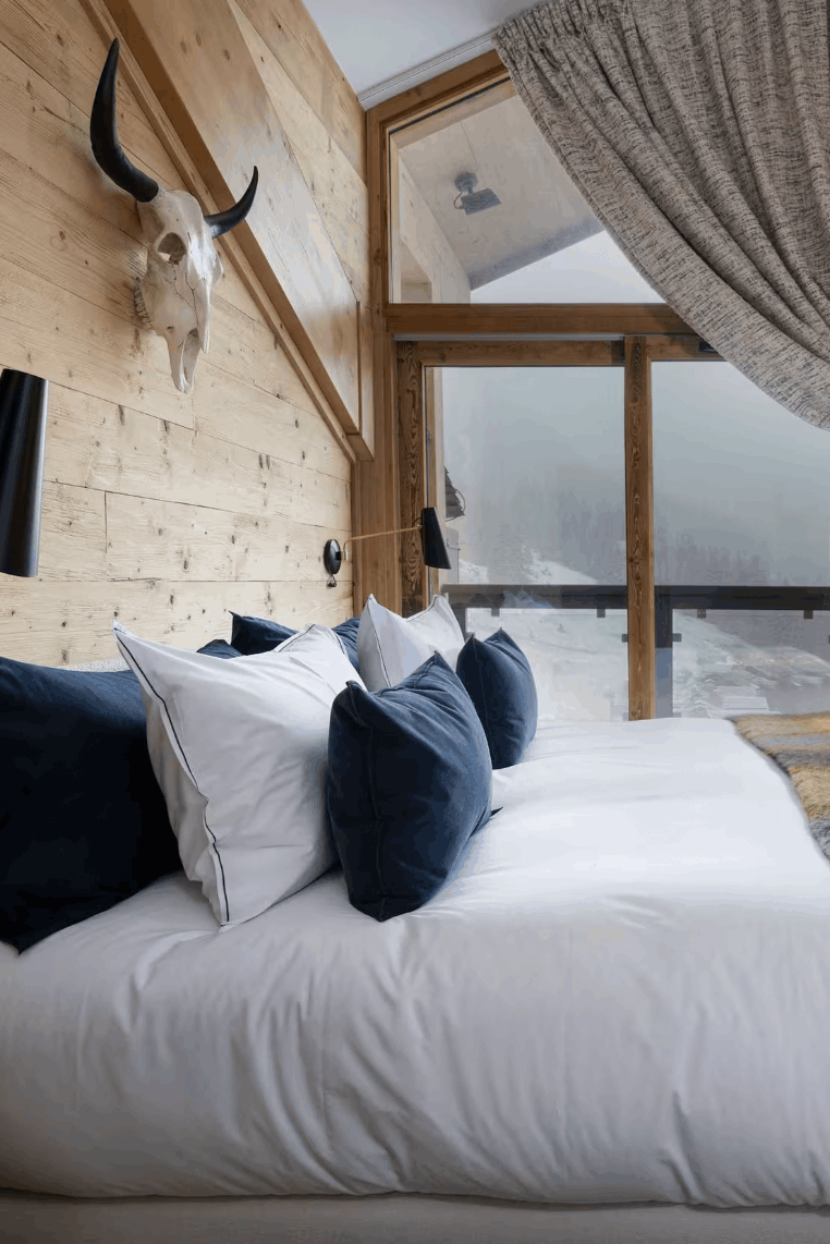 A modern bedroom interior with a large bed dressed in white bedding, dark navy and white pillows, and a yellow-and-brown plaid throw blanket. The room has light wood walls, floor-to-ceiling windows with gray curtains, and a mounted bull skull decor piece above the bed.