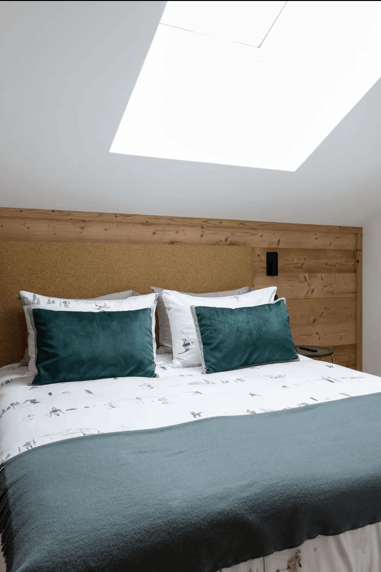 A modern bedroom featuring a bed dressed in white patterned bedding with two dark teal pillows and a dark teal throw blanket, set against a light wood headboard and matching wood sliding wardrobe doors.