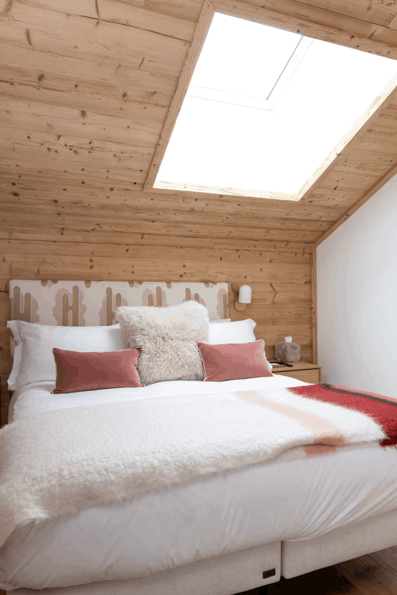 A cozy bedroom with a bed dressed in white bedding and red accent pillows, set in a rustic wooden attic room with a large skylight and white wall.