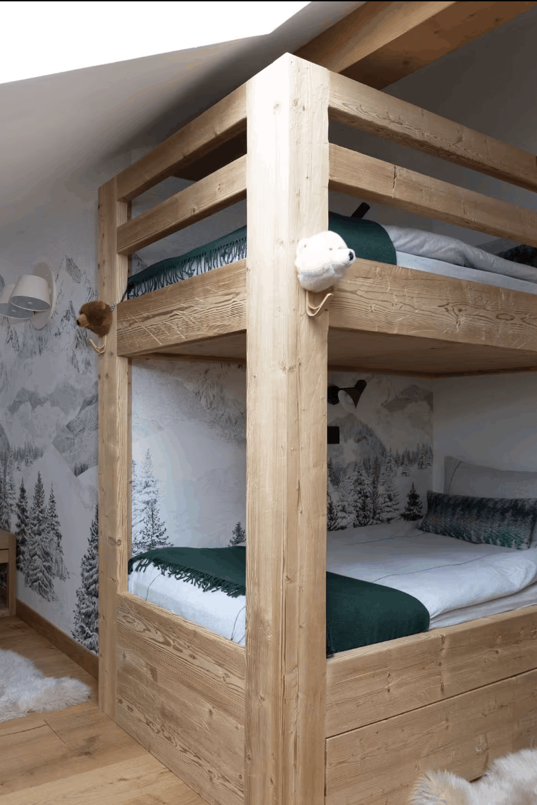 A rustic mountain-style bedroom featuring a light natural wood bunk bed with a ladder and a single single bed with a headboard, both styled with white and dark green bedding. The room has a slanted ceiling with a skylight, wood-paneled walls, and a forest-patterned accent wall.