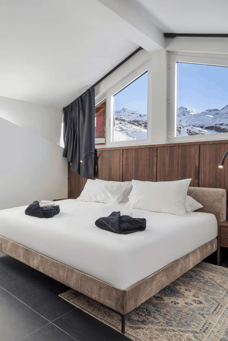 A modern ski resort bedroom featuring a double bed with white bedding, a wood-paneled headboard, large windows with a view of snowy mountains, a vanity with a marble countertop, and a patterned rug.