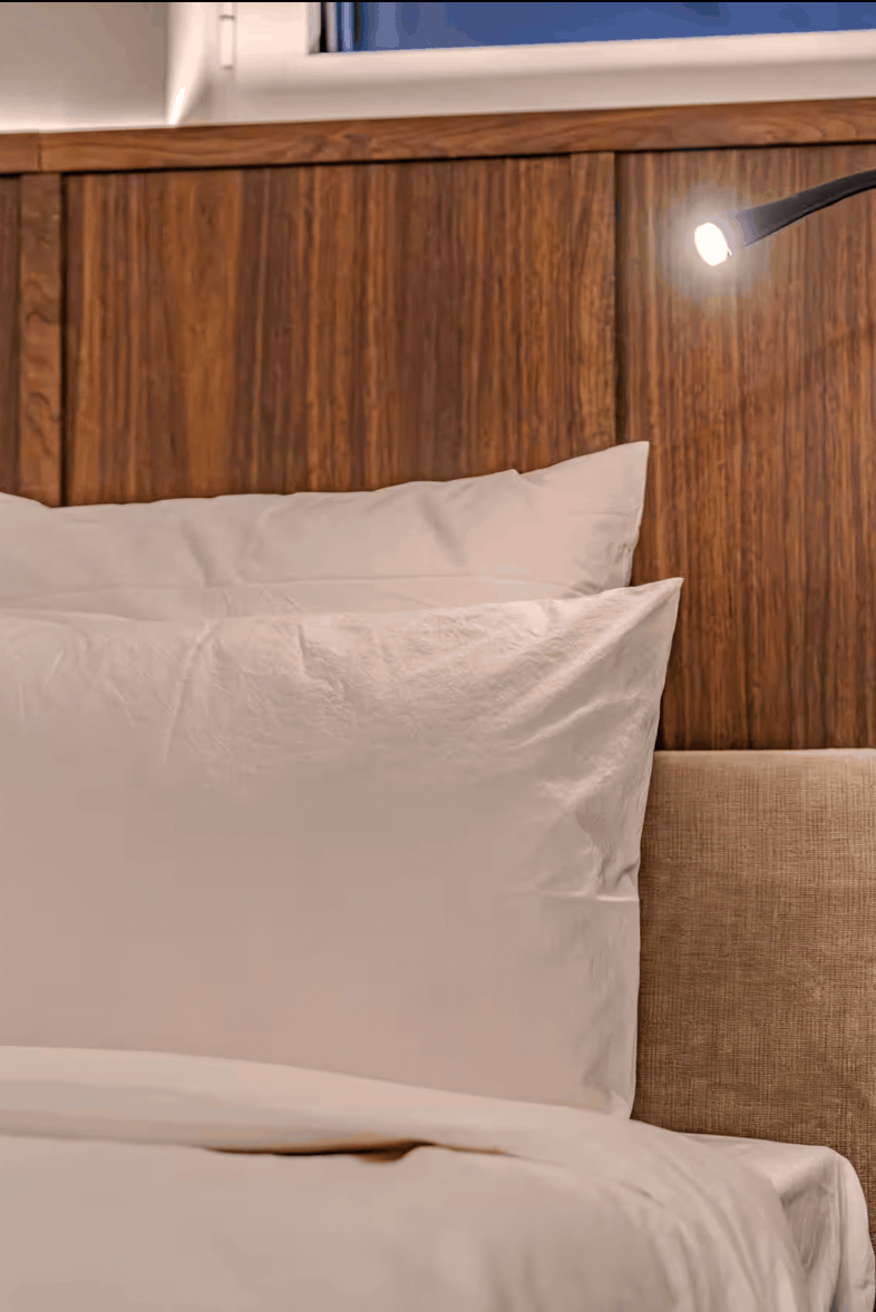 A close-up of a neatly made bed with white pillows and duvet, set against a dark wood-paneled headboard. A black adjustable reading light is mounted above, and a bedside table at right holds a water bottle and a dark glass cup.