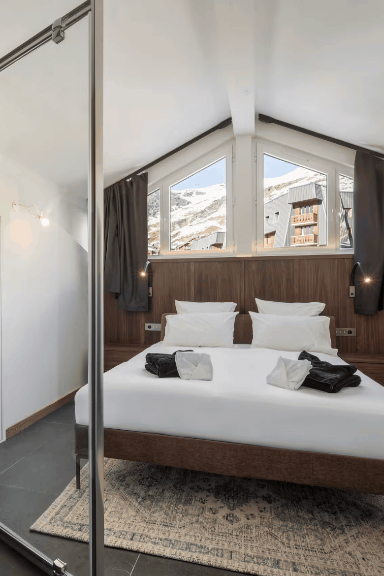 Modern minimalist bedroom with a large bed dressed in white linens, dark wood headboard and footboard, and a glass sliding door leading to a dark gray tiled shower area.