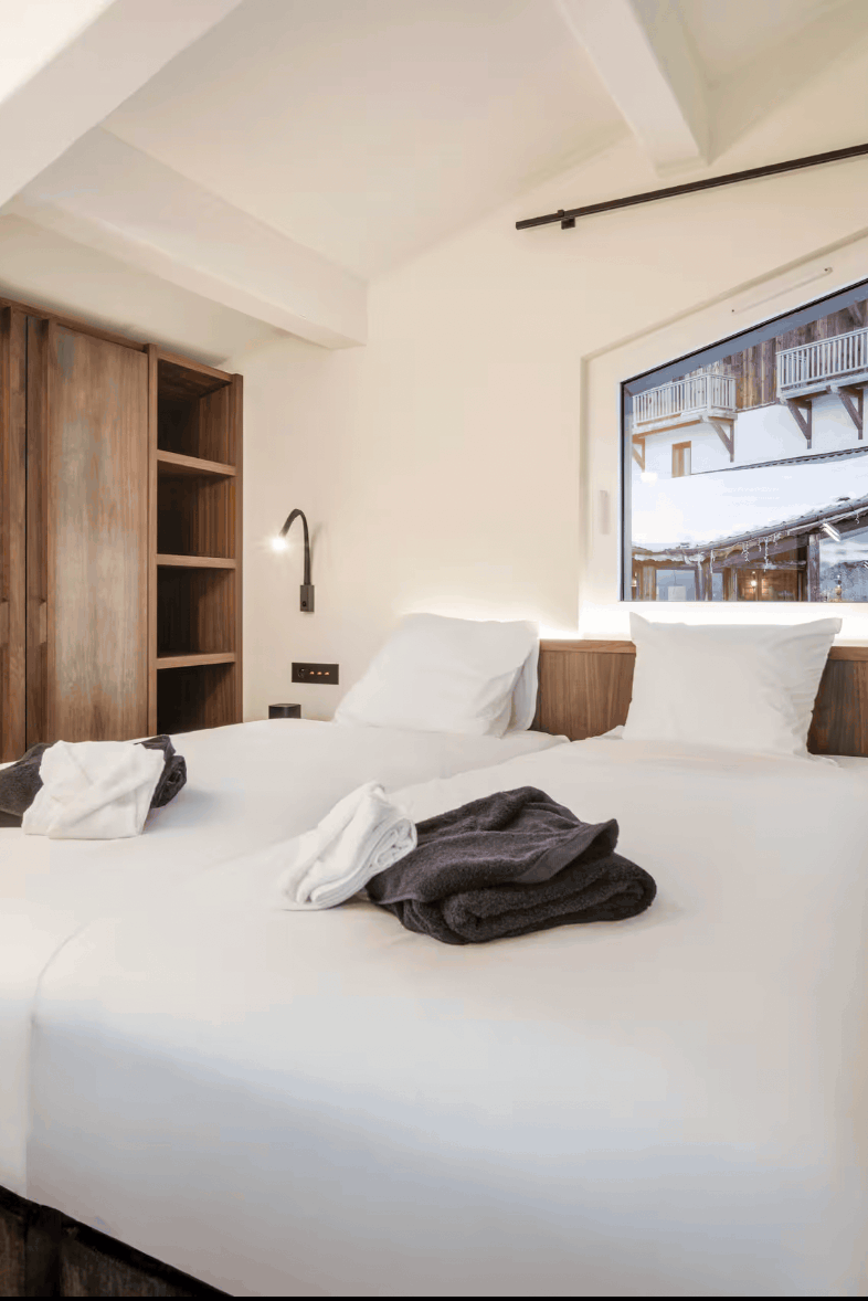 A modern bedroom interior featuring a neatly made bed with white linens, a large window with a view of snowy mountainside, dark gray curtains, wooden furniture, and a warm off-white wall.