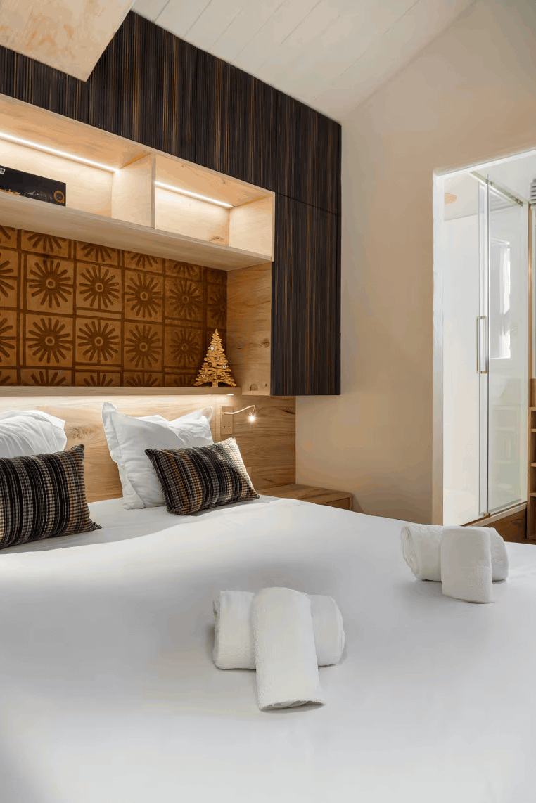 A modern bedroom interior featuring a large bed with white linens, striped pillows, built-in wooden shelving and headboard, warm ambient lighting, and a window looking out onto other buildings.