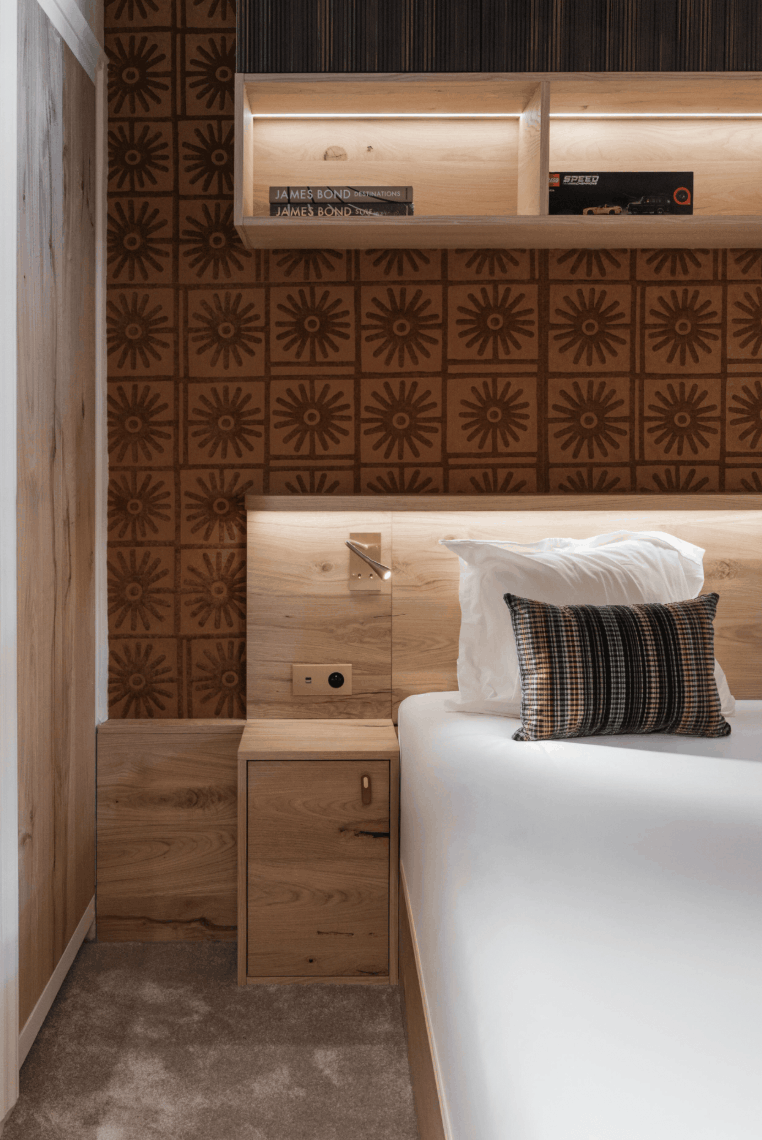 A modern bedroom corner featuring a bed with white bedding and a striped accent pillow, set against a wall with warm wood paneling, built-in shelves, and a patterned brown sunburst wallpaper.