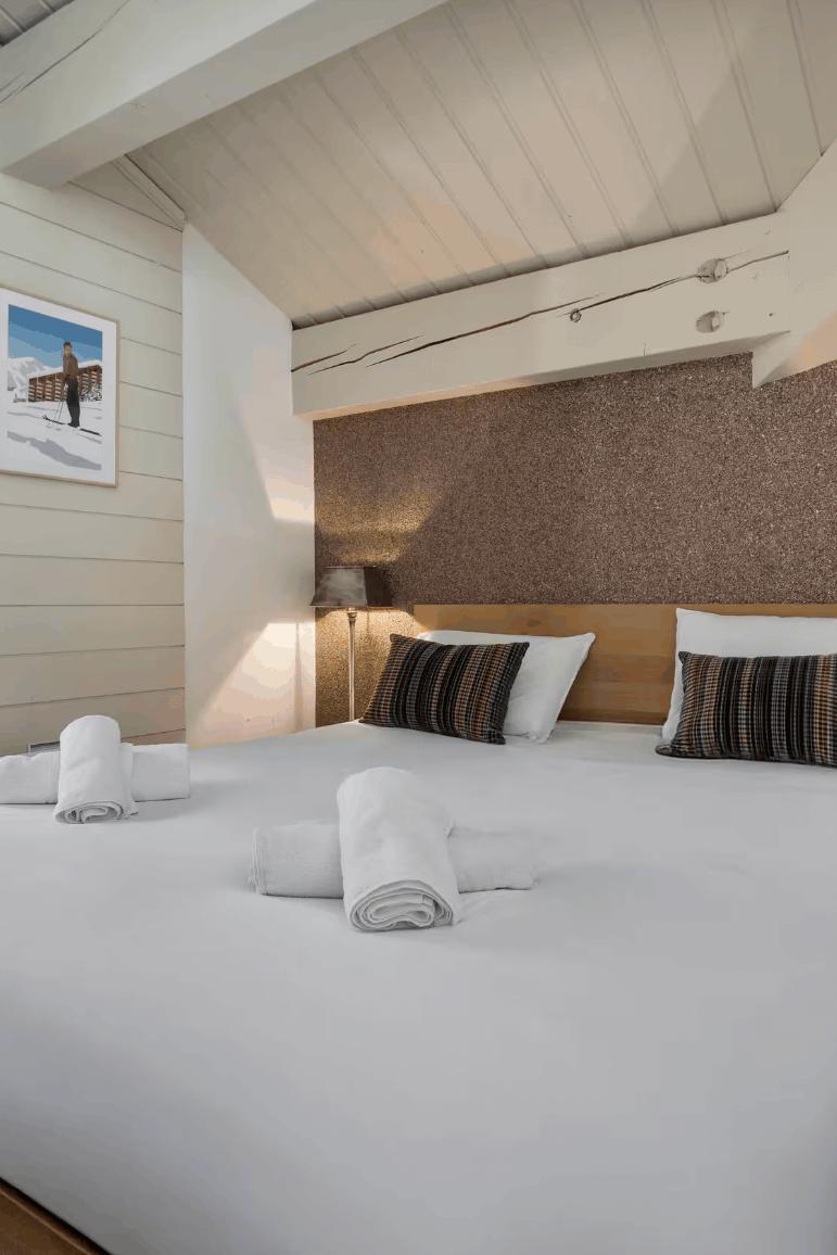 A modern bedroom interior with a large bed dressed in white linens, striped pillows, and rolled towels, set in a ski chalet-style room with wood ceiling beams, a textured brown accent wall, and a window overlooking a snowy outdoor scene.