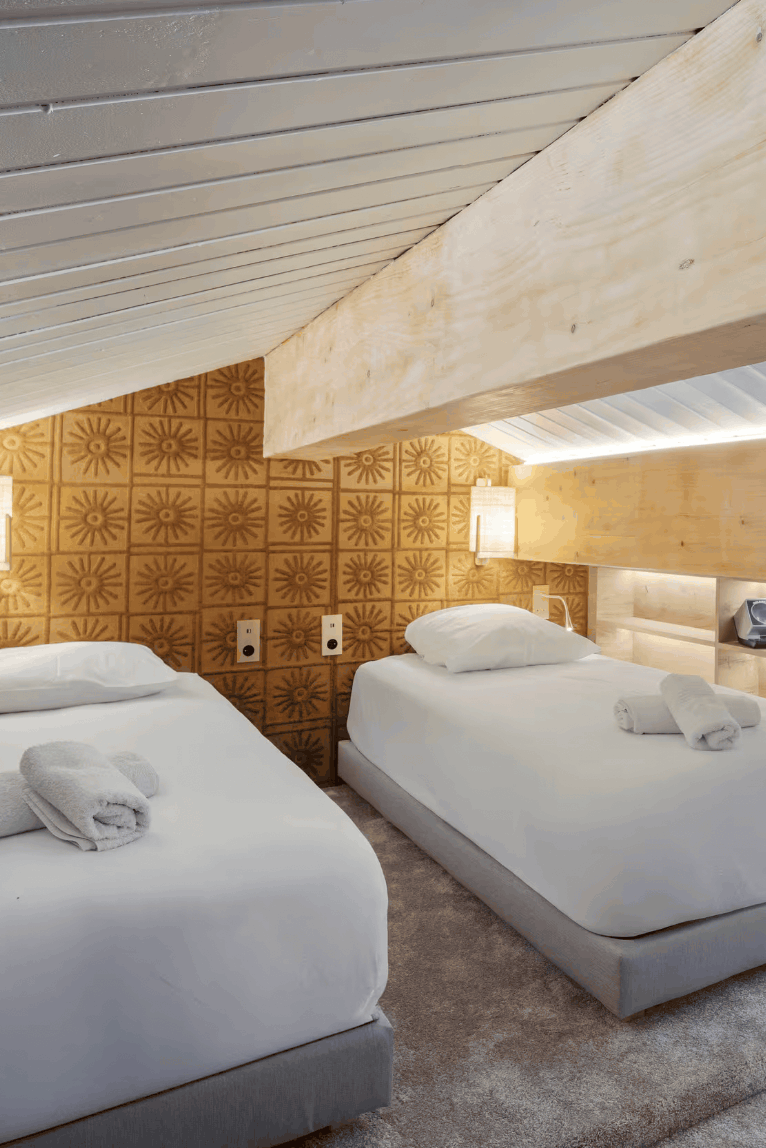 A modern rustic bedroom featuring two single beds with white linens, set under a slanted wooden ceiling with integrated shelves and warm ambient lighting.