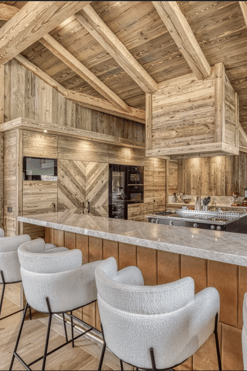 A luxury modern kitchen island in a rustic wooden chalet, featuring a marble countertop, white bar stools, a large wood range hood, and a high vaulted ceiling with exposed wooden beams.