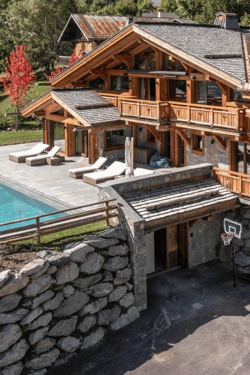 Aerial view of a large alpine-style chalet with wooden balconies, stone walls, a swimming pool, and a driveway, surrounded by trees and landscaping.