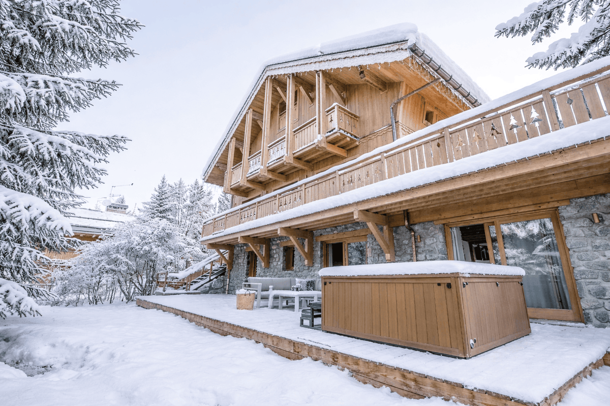 A snow-covered alpine chalet with wooden balconies, stone walls, and a wooden hot tub on a snowy patio, surrounded by snow-laden pine trees.
