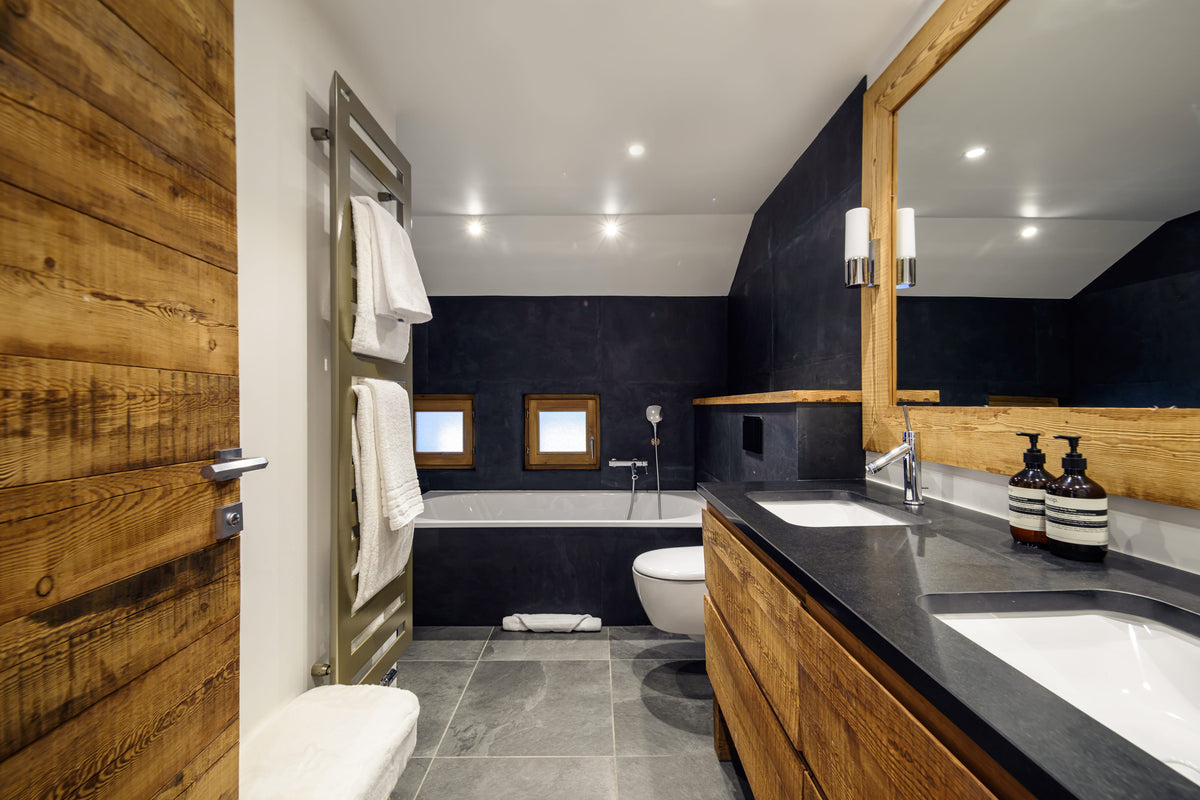 A modern luxury bathroom featuring a double vanity with a dark stone countertop and integrated sinks, rustic wood cabinets, a large wood-framed mirror, a heated towel rail, and a bathtub set against dark charcoal walls.