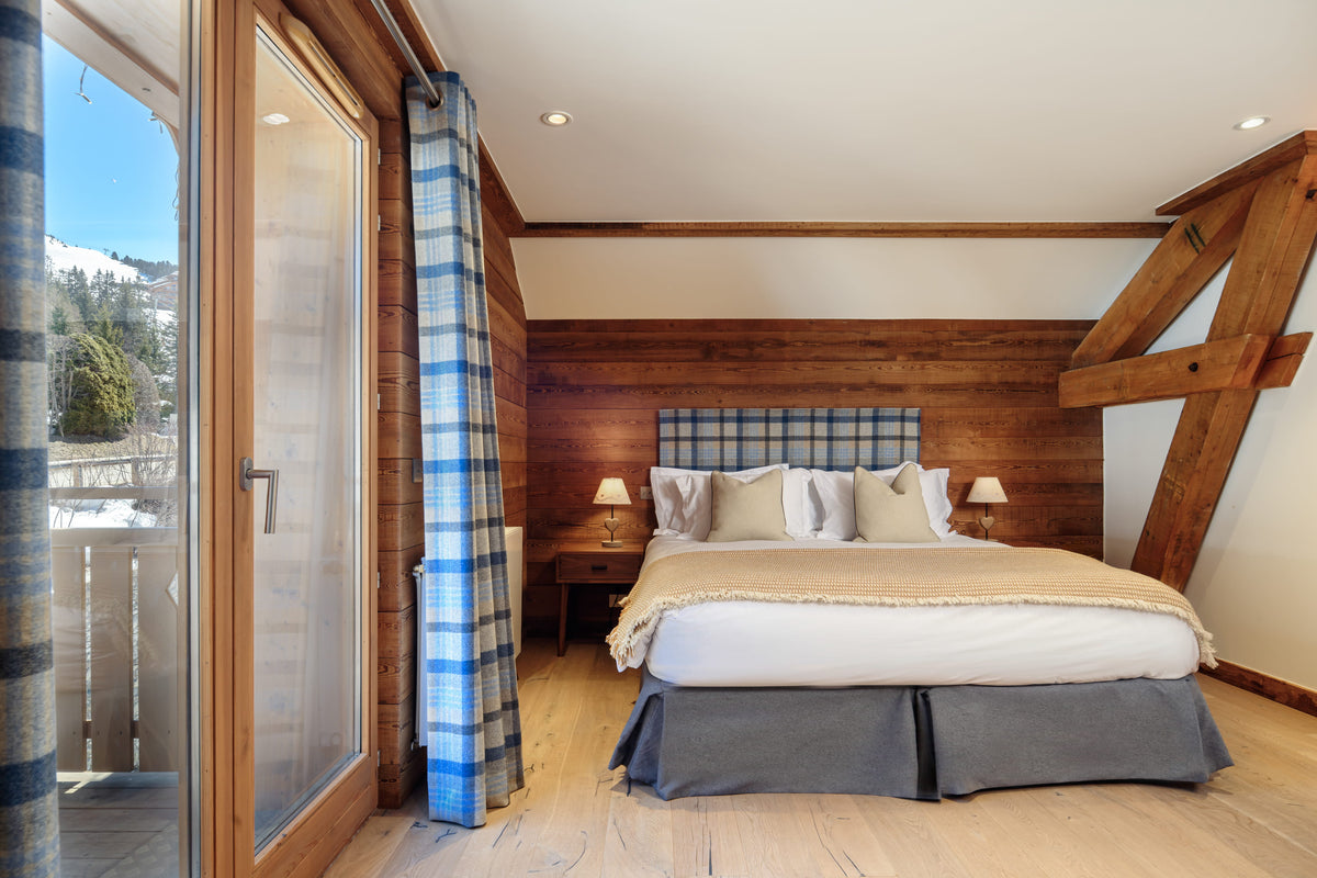 A rustic mountain chalet bedroom featuring a double bed with white bedding and gray skirt, set against a wood-paneled wall with a plaid headboard. Blue and white plaid curtains, light wood floors, and a large glass sliding door showing a snowy outdoor balcony are visible.