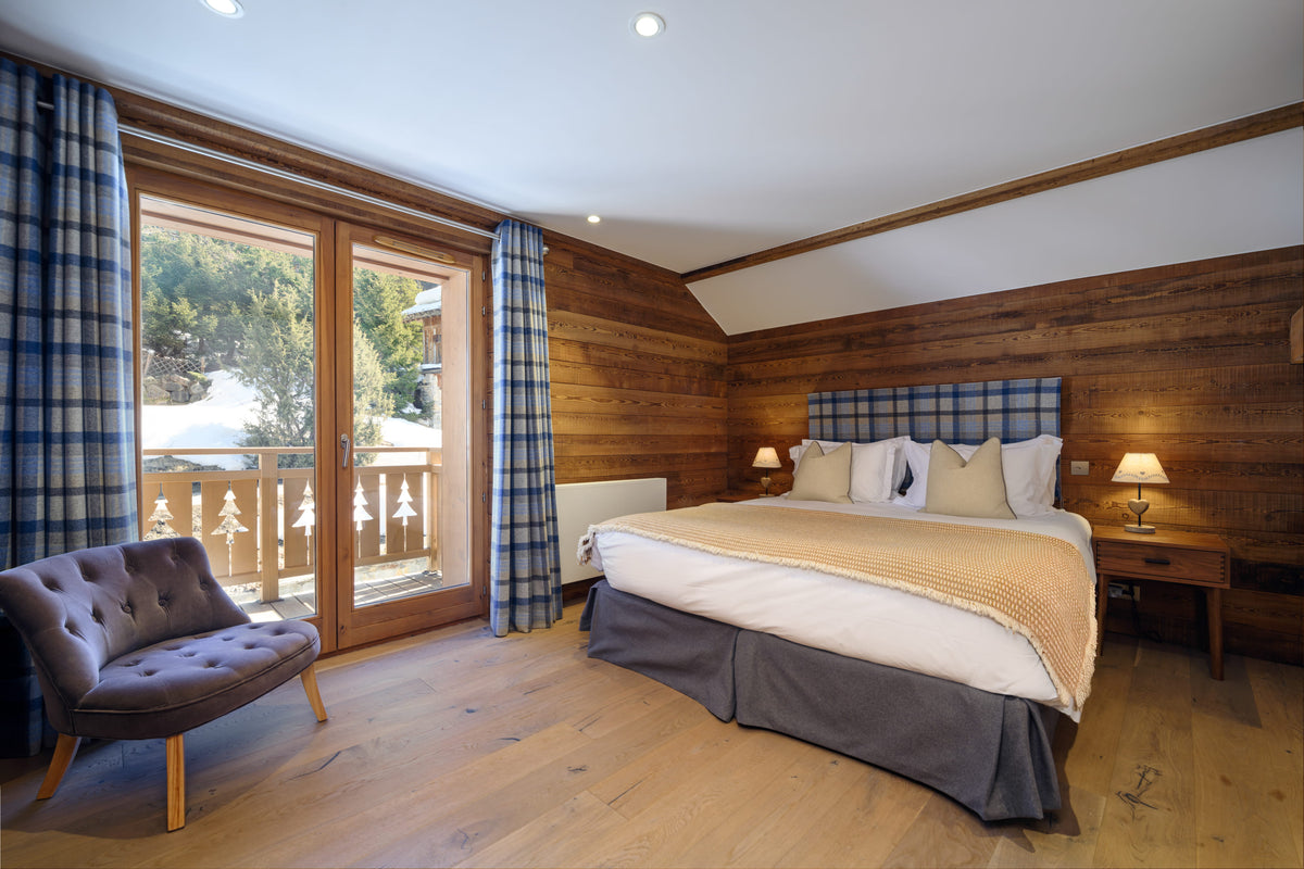 A cozy, modern ski chalet bedroom featuring a large bed with white and beige linens, a blue plaid headboard, wood-paneled walls, and a matching blue plaid curtain. A tufted gray armchair sits in the foreground near large glass doors opening to a balcony.
