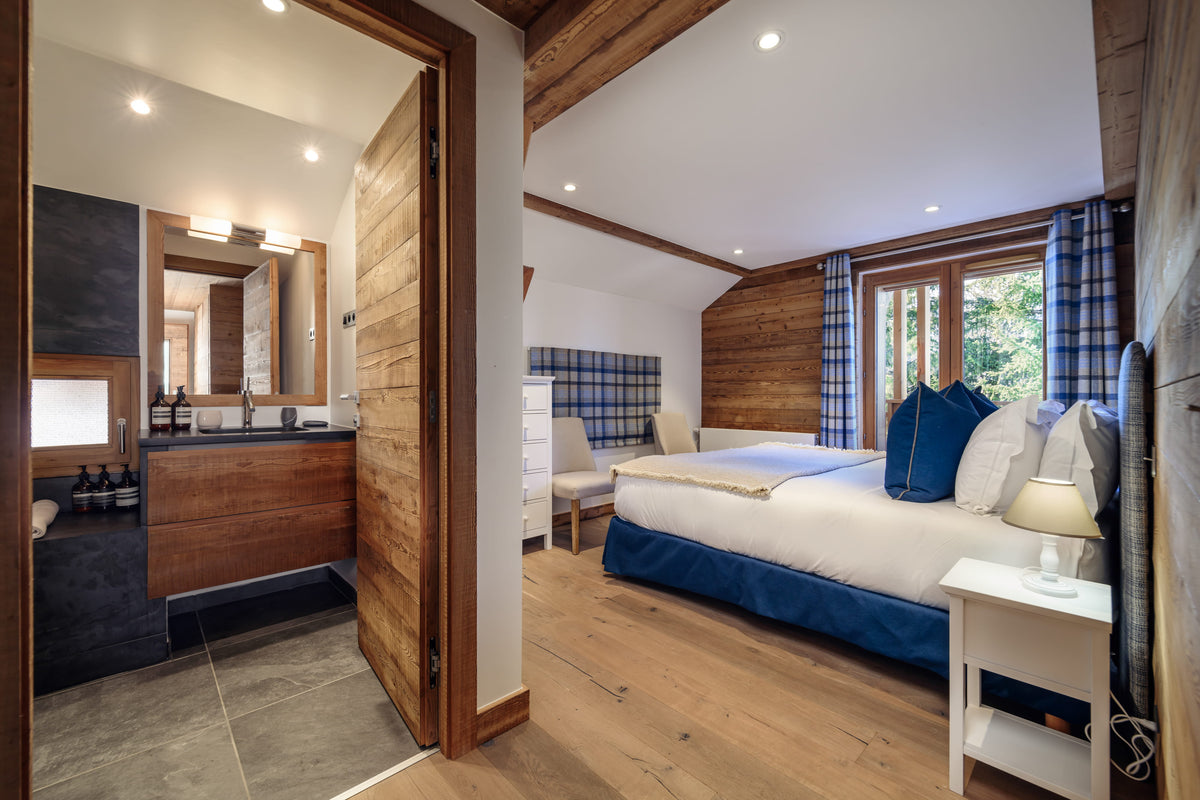 A modern ski chalet bedroom featuring a large bed with white bedding and blue accents, a matching blue plaid headboard and curtains, light wood floors, and an open doorway revealing a dark-tiled bathroom.
