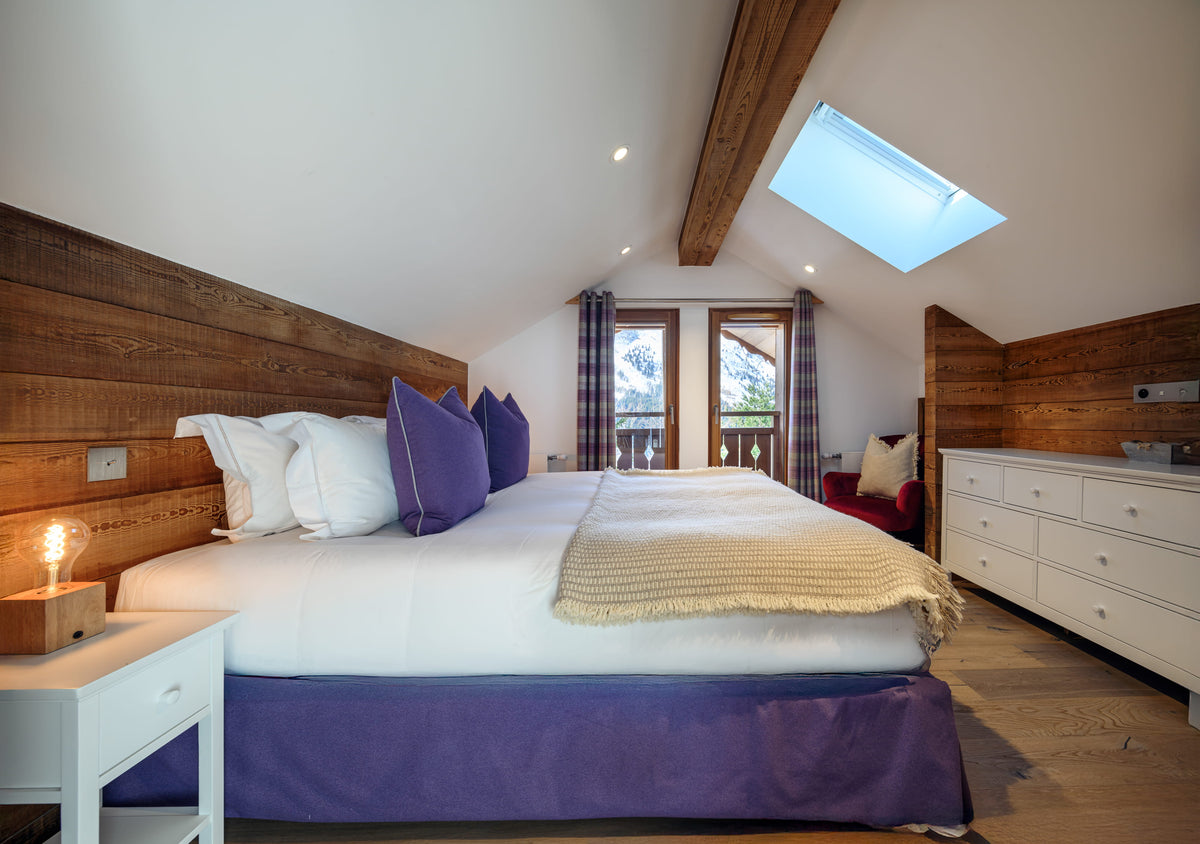 A modern bedroom with a large bed dressed in white bedding and purple pillows and bed skirt, set against a wood-paneled wall in an attic room with a skylight, white dresser, and wooden floor.