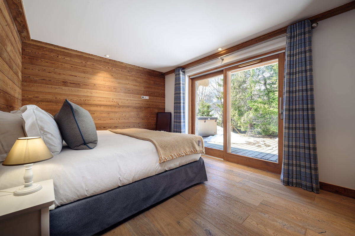 A modern bedroom with a bed dressed in white bedding and gray pillows, set against a wood-paneled wall. Large windows with blue plaid curtains look out to a garden patio.