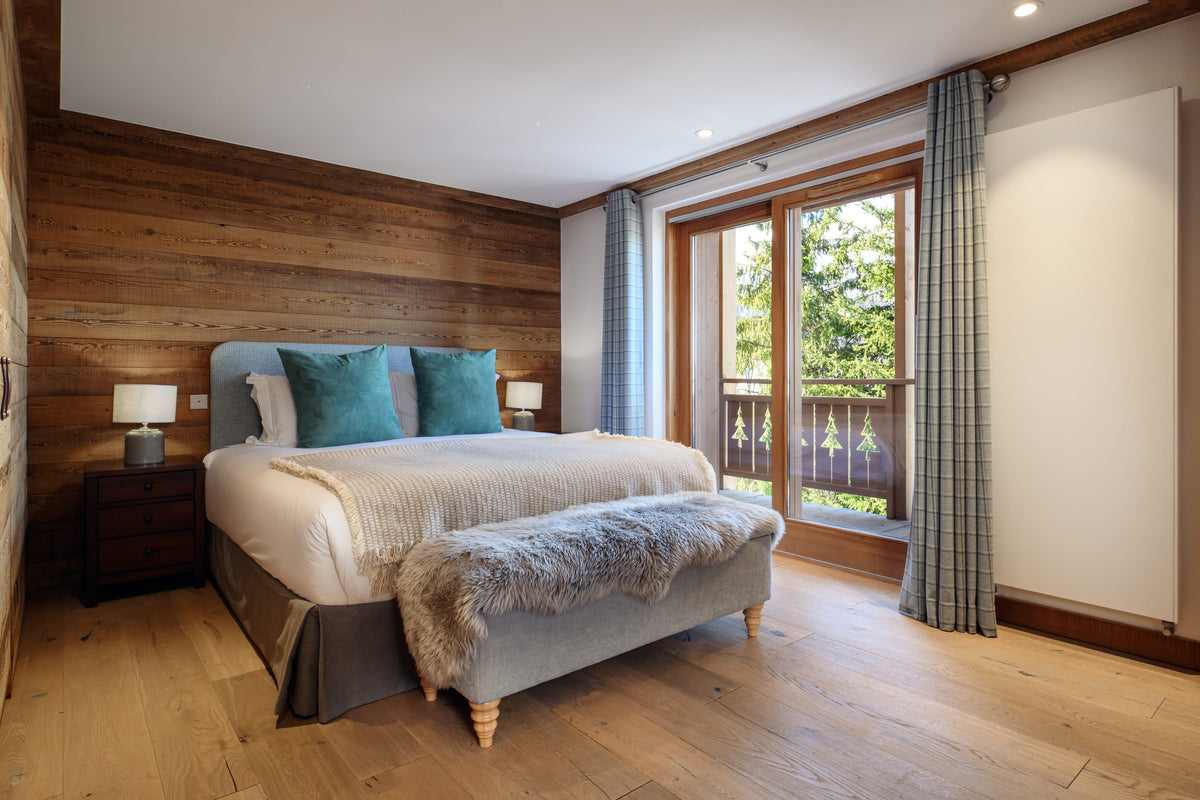 A modern bedroom featuring a double bed with white bedding and teal pillows, set against a wood-paneled wall. A gray upholstered bench with a faux-fur throw sits at the foot of the bed, and large windows with plaid curtains look out to a balcony and greenery.