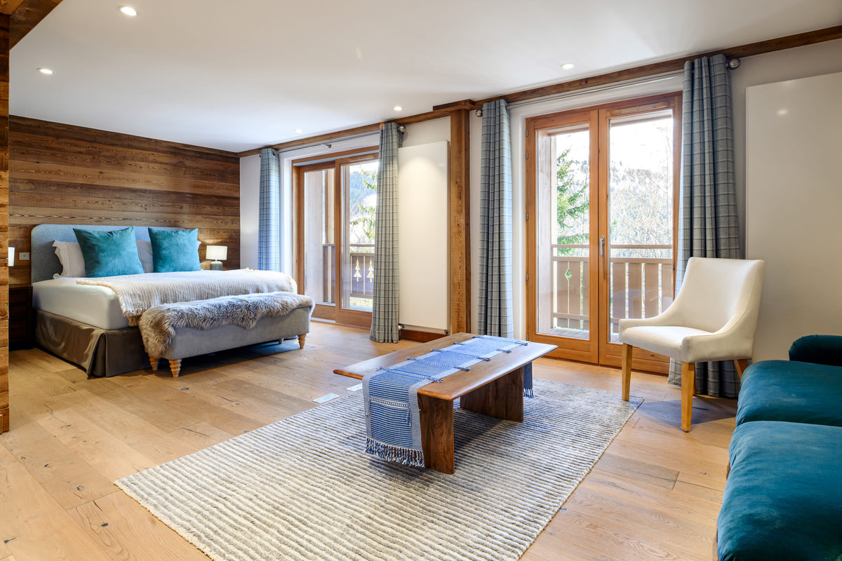 A modern ski chalet bedroom featuring a double bed with blue pillows, a wooden bench, a white armchair, and a blue sofa, set in a room with wood-paneled walls, large glass doors, and a striped rug.