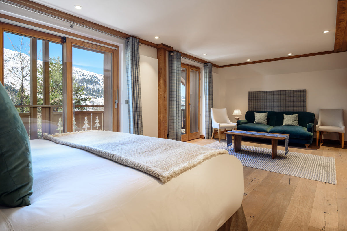 A luxury mountain chalet bedroom featuring a large bed in the foreground, a modern living area with a teal sofa, wooden floors, and floor-to-ceiling windows showcasing a snowy mountain landscape.