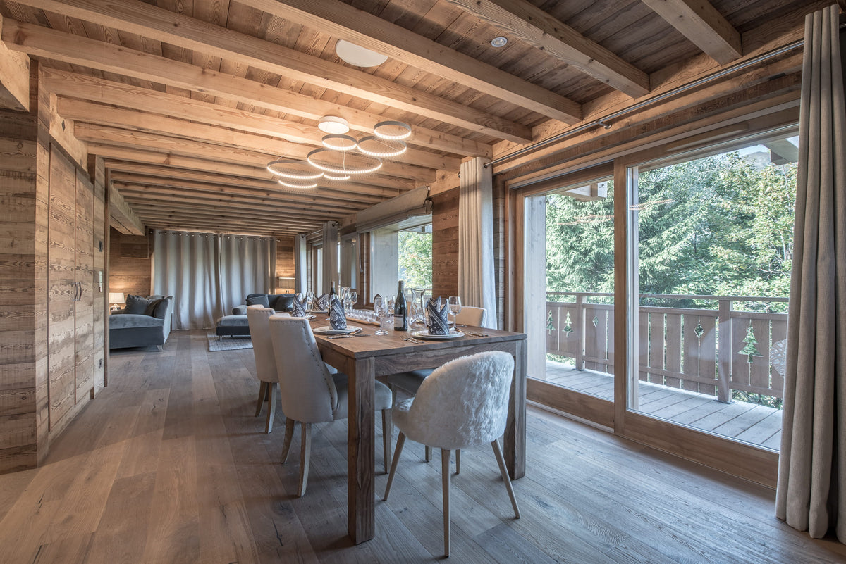 A modern dining room with a wooden table and textured white chairs, set in a rustic wooden chalet with large windows overlooking a green outdoor balcony.