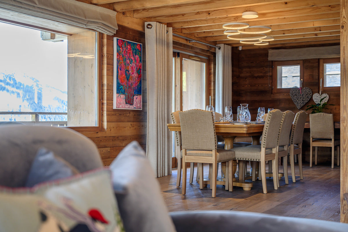 A rustic wooden dining room with a round wooden dining table and upholstered chairs, set in a chalet-style interior with wood-paneled walls and large windows showing a snowy mountain view.