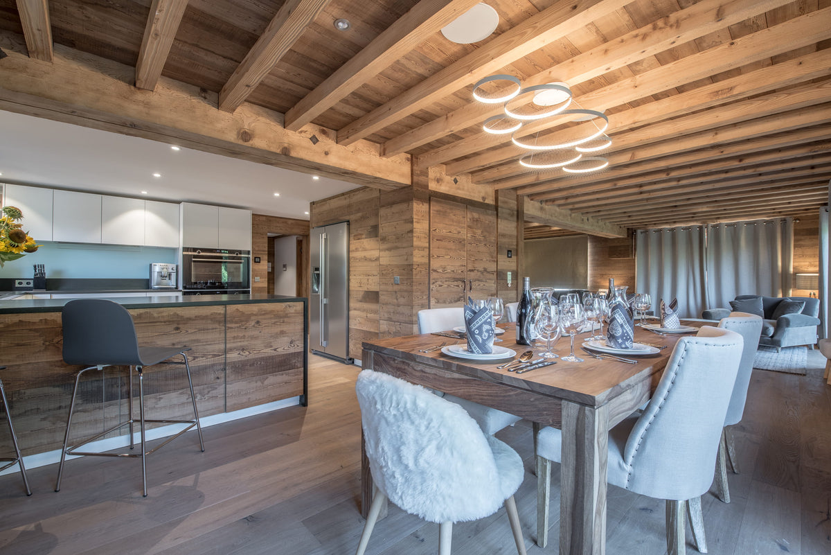 A luxury modern chalet interior featuring a rustic wooden dining room with a long wooden dining table, white textured chairs, a built-in wood kitchen island with a black bar stool, and a high ceiling with exposed wooden beams and circular pendant lights.