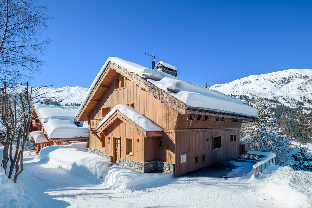 A rustic wooden chalet with snow-covered roofs sits in a snowy mountain landscape under a clear blue sky.
