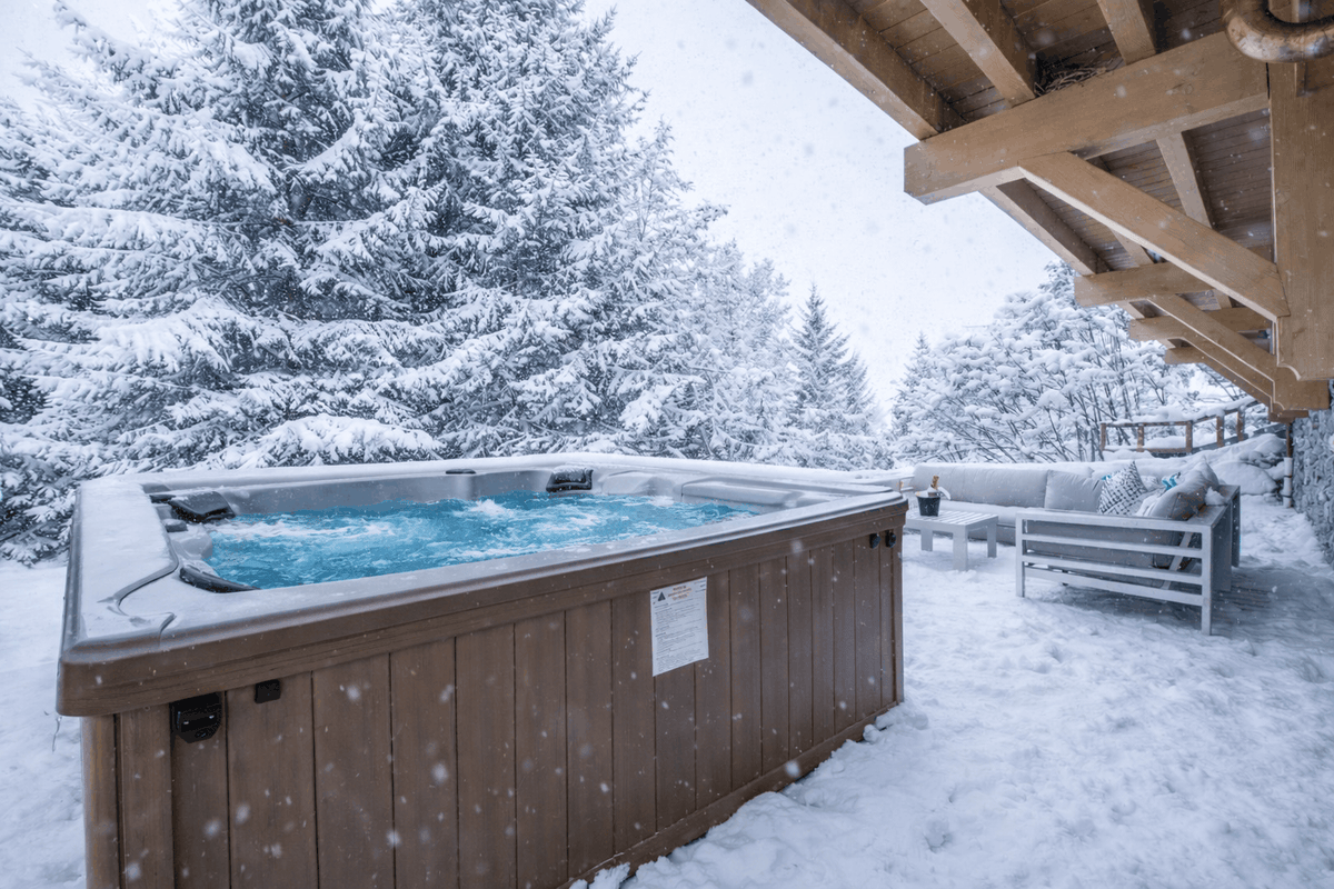 A wooden outdoor hot tub with blue water sits in a snowy backyard beside a covered patio, with snow-covered evergreen trees in the background.