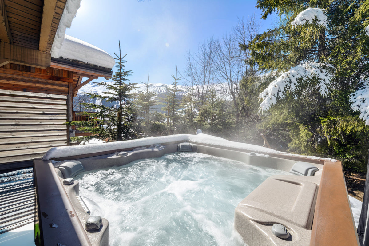 A hot tub with bubbling water sits outdoors on a snowy deck beside a wooden chalet, with snow-covered evergreen trees and mountains in the background under a blue sky.