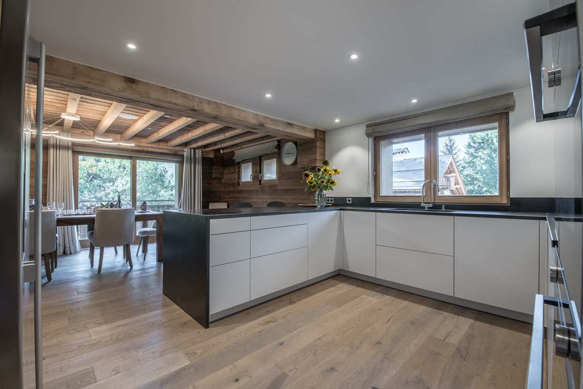 Modern kitchen interior with white lower cabinets, dark countertops, light wood flooring, and large windows. A dining area is visible through an open doorway on the left.