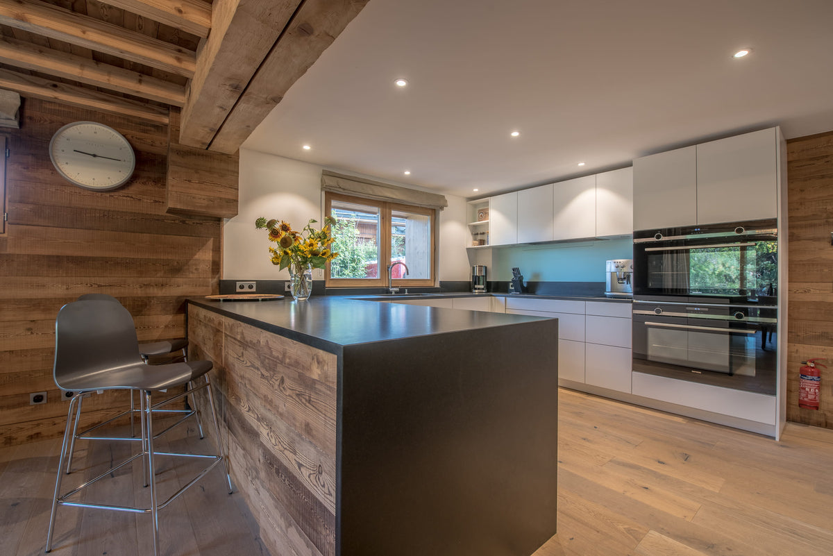 A modern kitchen interior with a large wood-and-metal island, white cabinetry, built-in stainless steel appliances, light wood flooring, and exposed wooden ceiling beams.