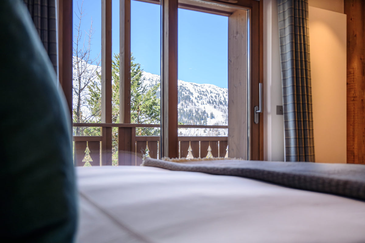 A close-up view of a bed with white bedding and a gray textured runner, positioned in front of large wooden-framed windows that offer a scenic view of snow-covered mountains and pine trees under a clear blue sky.