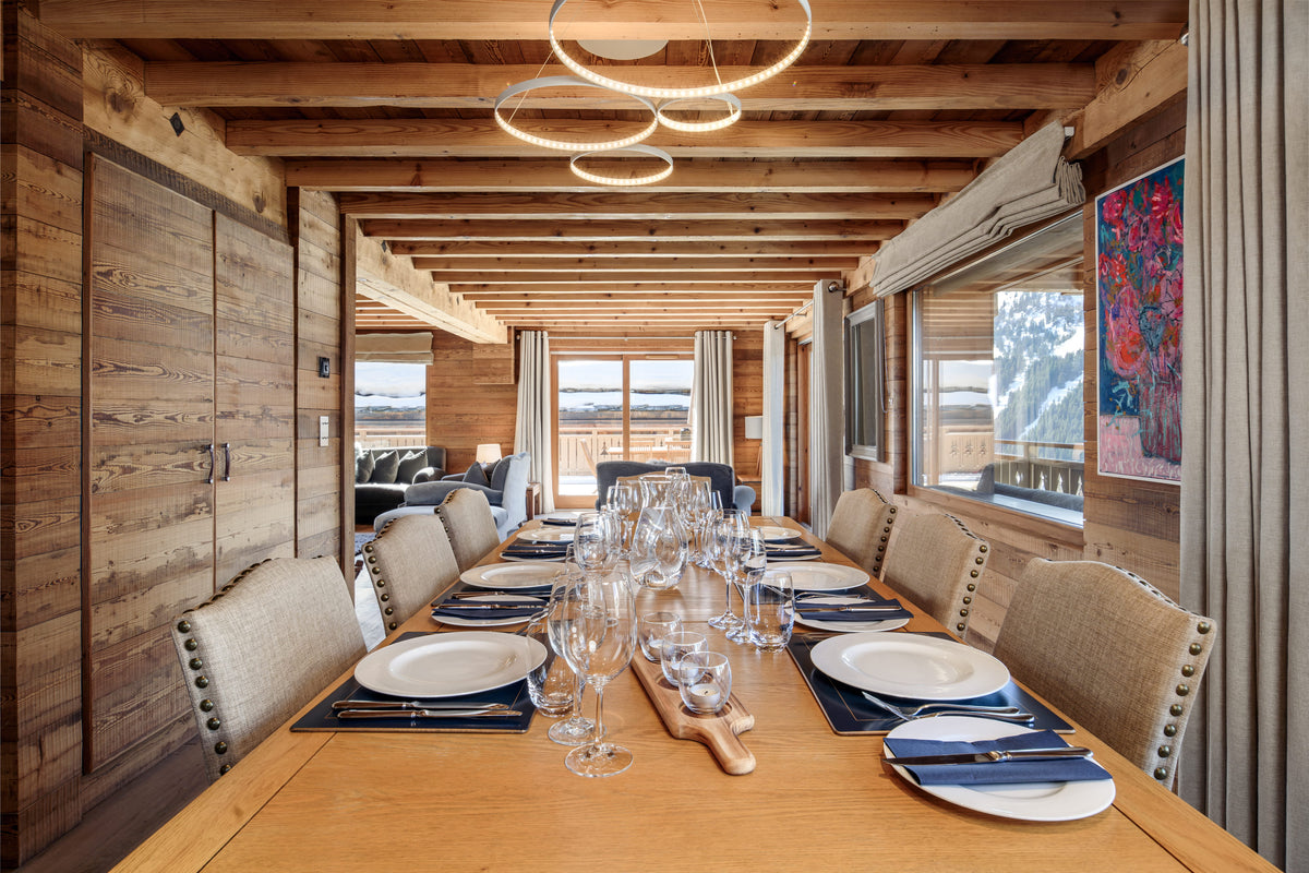 A long wooden dining table set with white plates, blue napkins, and clear wine glasses inside a rustic chalet with wood-paneled walls and ceiling.