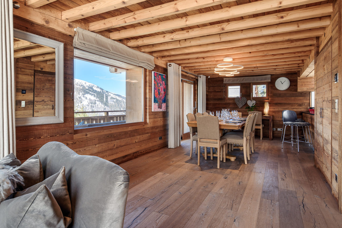 A rustic wooden chalet interior with exposed wood ceiling beams, hardwood floors, a dining table and chairs, a gray sofa in the foreground, and a large window showing a snowy mountain view.