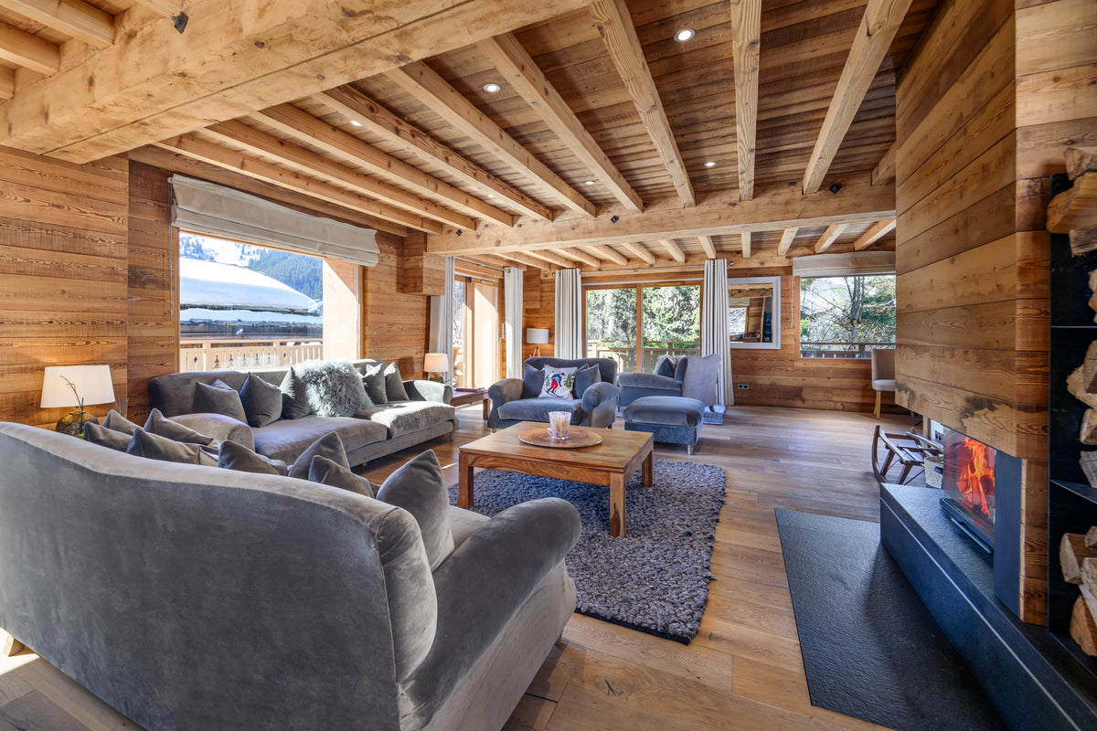A luxurious mountain chalet living room featuring a large gray velvet armchair in the foreground, a matching gray sectional sofa, a wooden coffee table, and a blue fireplace. The room is characterized by rustic wooden walls, exposed wooden ceiling beams, and large windows looking out onto snowy mountains.
