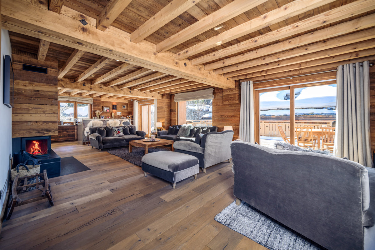 A spacious, rustic mountain chalet living room with exposed wood ceiling beams, hardwood floors, a black wood-burning fireplace, and modern gray sofas with an outdoor deck visible through large windows.