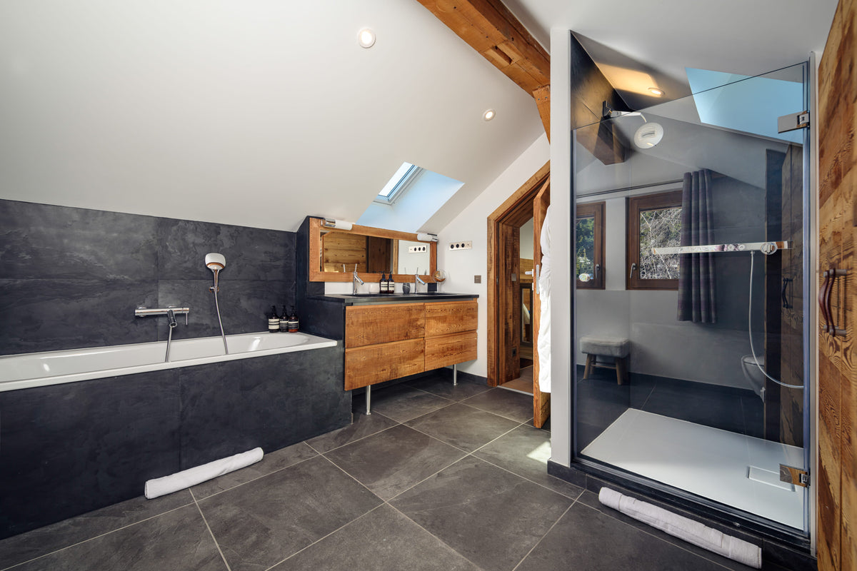A luxury modern bathroom featuring a large white bathtub set into a dark gray stone wall, a matching wood vanity, and a glass-enclosed walk-in shower with a white walk-in tray. The room has a high vaulted ceiling with exposed wooden beams and skylights.