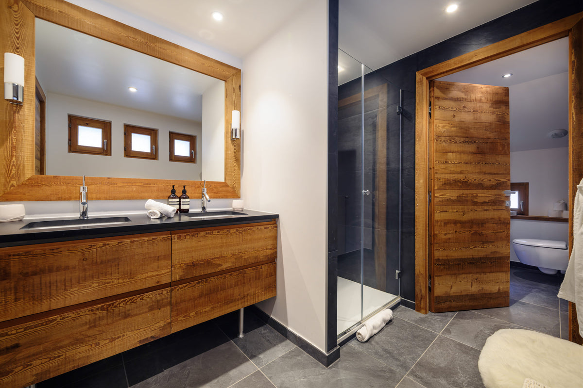 A modern bathroom interior featuring a large wood-framed double vanity with a black countertop and sink, a glass shower enclosure, dark gray floor tiles, and an open wooden door leading to a bathtub.