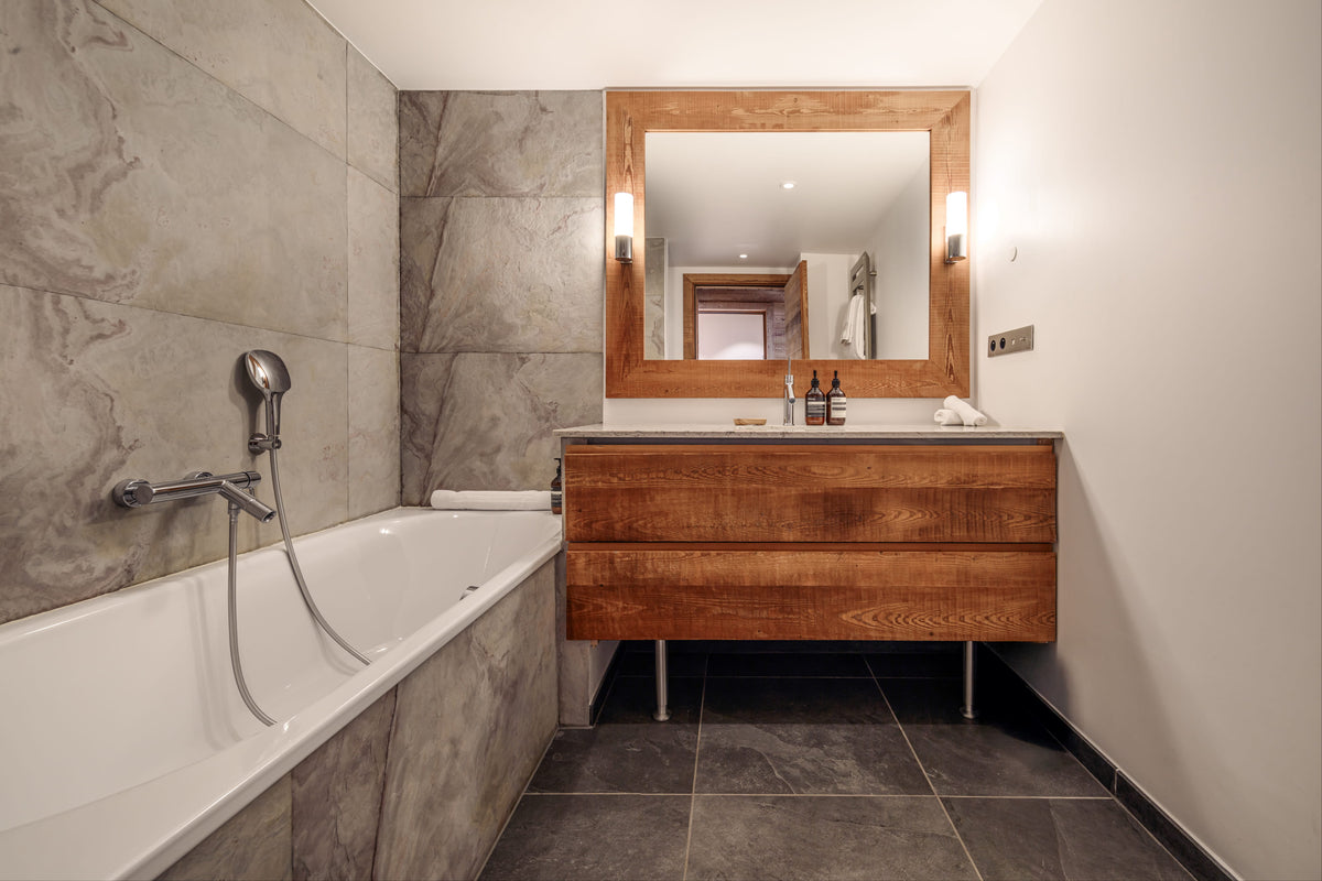 A modern bathroom interior featuring a white bathtub with a chrome handheld shower, a floating wooden vanity with a matching wood-framed mirror, and dark gray stone tile flooring and walls.