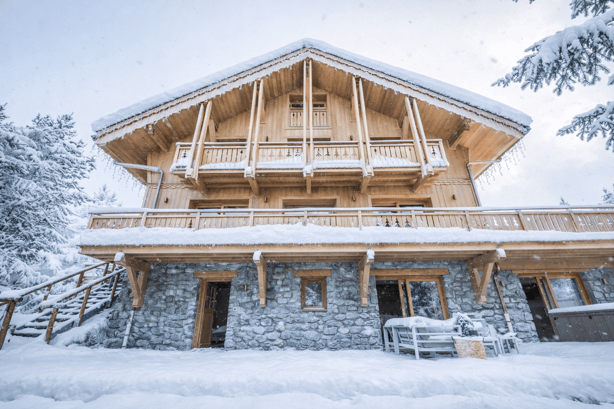 A large alpine-style wooden chalet with snow-covered balconies and stone lower walls, surrounded by deep snow and evergreen trees in a winter landscape.